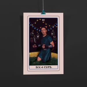 May include: A tarot card print featuring a person in a teal outfit and cat ears, standing in a tub of green liquid. The card is labeled "SIX of CUPS" and has the Roman numeral "VI" at the top. Decorative bottles and string lights are in the background.