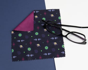 May include: A dark blue microfiber cleaning cloth with a repeating pattern of green eyes, planets, stars, and wings. The cloth is folded in half and has a burgundy colored side. A pair of black framed glasses is lying on top of the cloth.