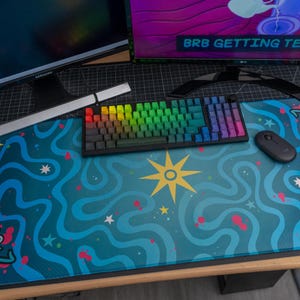 May include: A large rectangular desk mat featuring a vibrant teal and blue wavy pattern with a central yellow star, small white stars, pink accents, and cartoonish blue creatures. The mat sits under a rainbow-colored mechanical keyboard and a black mouse.