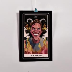 Puede incluir: Un póster de tarot que representa "El Diablo". Muestra una figura sonriente con cuernos, rodeada de llamas. La carta está enmarcada en negro con el número romano "XV" en la parte superior y el título en la parte inferior.