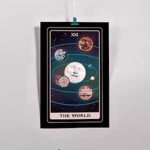 May include: A framed tarot card print featuring a cosmic design. The card depicts a large white moon with a face, surrounded by planets and swirling blue lines against a dark background. The text "THE WORLD" is at the bottom.
