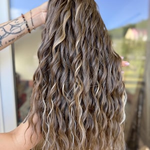 May include: A long, wavy wig with a mix of brown, blonde, and light brown highlights. The hair is styled in loose, flowing curls. The wig is held up, showcasing the length and texture of the hair.