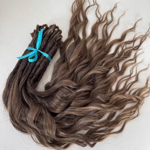 Puede incluir: Un conjunto de rastas de color marrón oscuro atadas con una cinta turquesa, junto a una sección de cabello largo y ondulado de color marrón claro con reflejos. El cabello está peinado para que parezca natural y fluido, con una mezcla de tonos oscuros y claros.