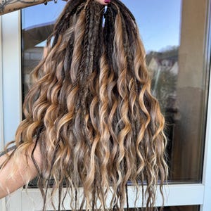 Op de afbeelding: Een set bruine en blonde dreadlocks met een golvende textuur. De dreadlocks worden tegen een raam gehouden, waarbij de verschillende tinten bruin en blond worden getoond. De haarverlengingen hebben een natuurlijke, getextureerde uitstraling.