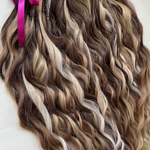 May include: A bundle of wavy, multi-toned dreadlocks in shades of brown, blonde, and white, tied with a bright pink ribbon. The hair extensions have a natural, textured appearance, suitable for various hairstyles.