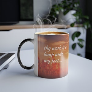 May include: A white coffee mug with a black handle. The mug has a heat-sensitive design that reveals a sunset scene with palm trees and the text "thy word is a lamp unto my feet..." when hot liquid is poured inside.