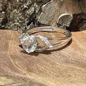 May include: A silver ring with a large, clear, round gemstone as the focal point. The ring features a split band design with smaller accent stones on either side of the main stone. The ring is set against a natural wood background.