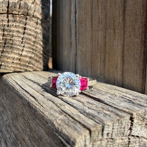 May include: A silver ring with a large, round, clear center stone flanked by two smaller, rectangular, red stones. The ring is set on a weathered wooden surface, with the sunlight highlighting the ring's details.