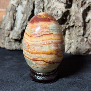May include: A polished, egg-shaped stone sculpture featuring horizontal bands of red, orange, yellow, and cream colors. The smooth, rounded object is displayed upright on a small, circular, dark brown wooden stand, showcasing natural mineral layers.