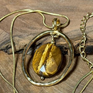 May include: A gold-toned necklace featuring a thin snake chain and a pendant composed of an oval-shaped, faceted tiger's eye gemstone suspended within a larger, smooth, circular gold-toned metal frame that hangs from a small metal bail.