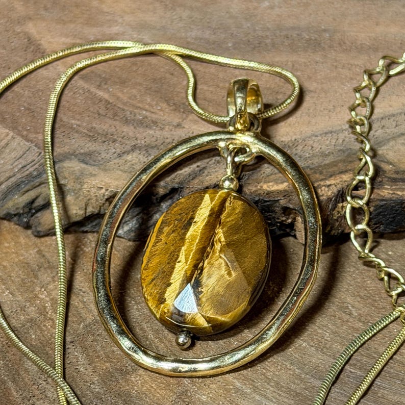 May include: A gold-toned necklace featuring a thin snake chain and a pendant composed of an oval-shaped, faceted tiger's eye gemstone suspended within a larger, smooth, circular gold-toned metal frame that hangs from a small metal bail.