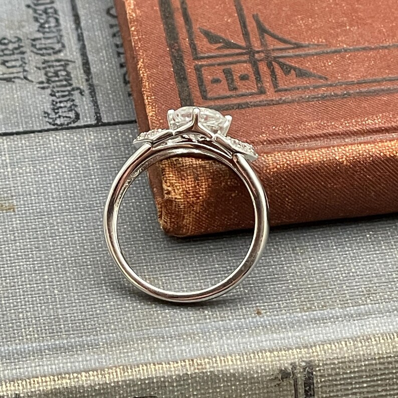 May include: A silver engagement ring with a large, round, clear gemstone as the centerpiece. The ring has smaller accent stones on either side of the main stone. The ring is set against a backdrop of a vintage book.