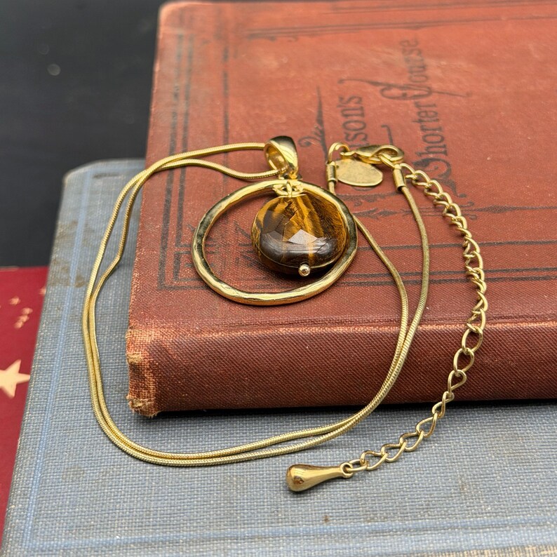 May include: A gold-tone necklace featuring a round, faceted tiger's eye gemstone pendant suspended within a circular gold-tone frame. The chain is a thin snake style with an adjustable extender chain ending in a small, smooth teardrop-shaped charm.