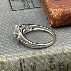 May include: A silver ring with a large, clear, square-cut stone as the focal point. The ring has a split shank design and is accented with smaller stones. The band is marked with "925". The ring is set on a textured, gray surface.