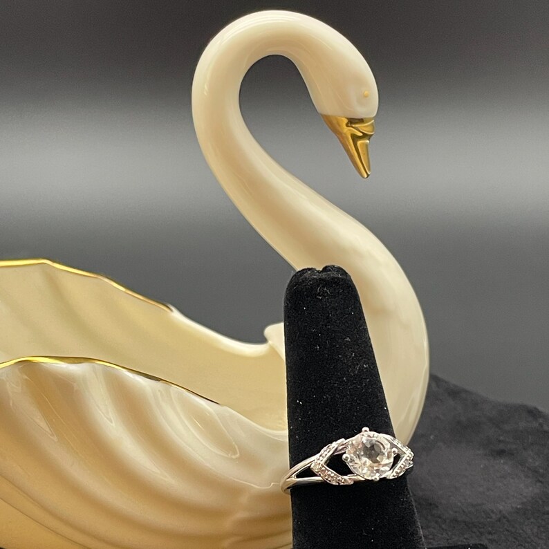 May include: A silver ring with a large, round, clear gemstone and smaller accent stones. The ring is displayed on a black ring holder in front of a white swan figurine with gold accents.