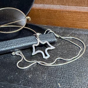 May include: A silver-toned necklace with a pendant in the shape of a stylized star or abstract figure. The necklace is on a black surface, with a pair of gold-rimmed eyeglasses in the background.