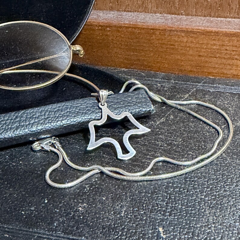 May include: A silver-toned necklace with a pendant in the shape of a stylized star or abstract figure. The necklace is on a black surface, with a pair of gold-rimmed eyeglasses in the background.
