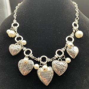 May include: Silver-toned necklace featuring heart-shaped pendants with ornate designs and faux pearls. One heart pendant has the inscription "Love the Lord your God with all your heart Luke 10:27". The necklace is displayed on a black surface.