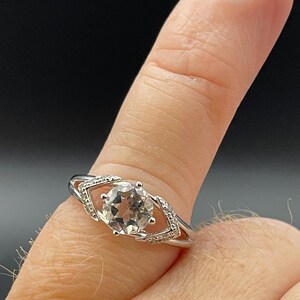 May include: A silver ring with a large, round, clear gemstone as the focal point. The ring features a decorative setting with smaller accent stones and a split band design. The ring is shown on a finger against a dark background.
