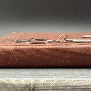 May include: A silver-toned necklace with a pendant shaped like a stylized wrench. The pendant is resting on a vintage, reddish-brown book. The necklace chain is a thin, flexible, silver-colored snake chain.