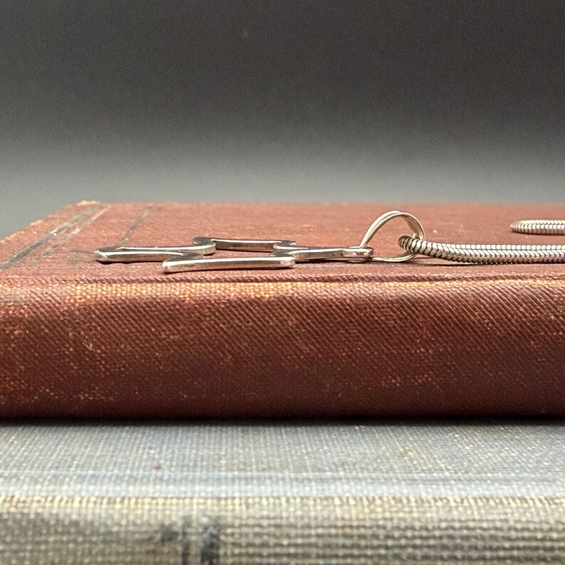 May include: A silver-toned necklace with a pendant shaped like a stylized wrench. The pendant is resting on a vintage, reddish-brown book. The necklace chain is a thin, flexible, silver-colored snake chain.
