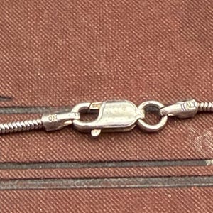 May include: Close-up of a silver necklace clasp with a snake chain. The clasp is marked with "925" and "Italy". The background is a textured brown surface.