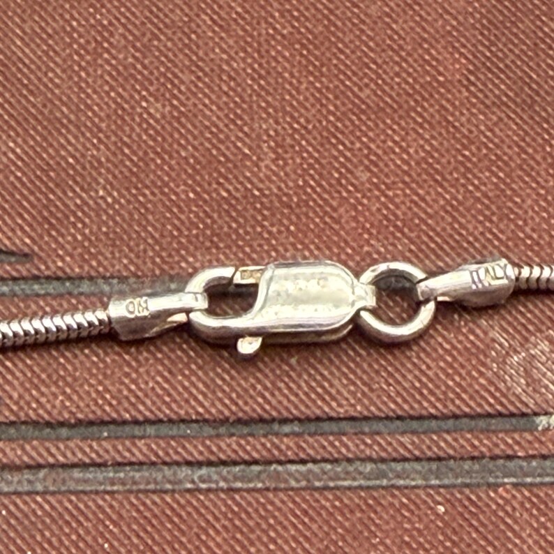 May include: Close-up of a silver necklace clasp with a snake chain. The clasp is marked with "925" and "Italy". The background is a textured brown surface.