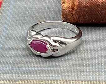 May include: A silver ring with a marquise-cut, deep red gemstone. The ring has a decorative, flowing design around the gemstone. The ring is set on a textured, gray surface.