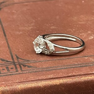 May include: A silver ring with a large, oval-shaped, clear gemstone as the centerpiece. The ring features a split band design and smaller accent stones on the sides of the main stone. The ring is set on a brown surface.