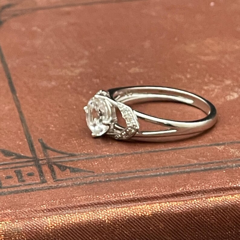 May include: A silver ring with a large, oval-shaped, clear gemstone as the centerpiece. The ring features a split band design and smaller accent stones on the sides of the main stone. The ring is set on a brown surface.