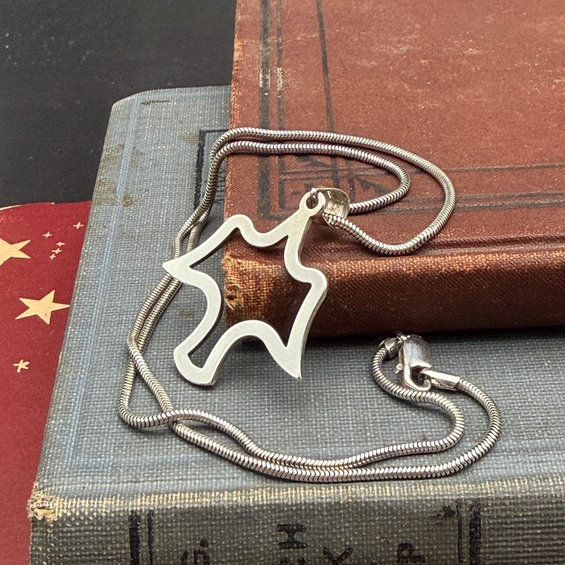 May include: A silver-colored necklace with a pendant in the shape of a stylized bear. The necklace chain is a thin, snake-like design. The pendant is a cutout design. The necklace is resting on a stack of vintage books.