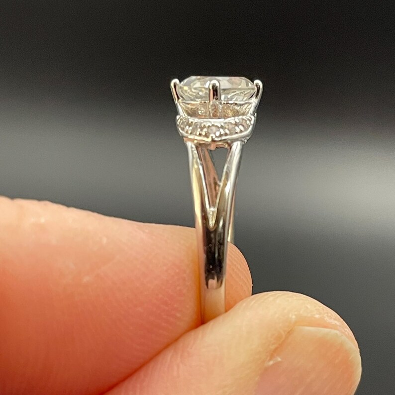 May include: A silver ring with a large, round, clear gemstone. The ring has a halo of smaller stones around the main stone and a split shank design. The ring is held against a dark background.