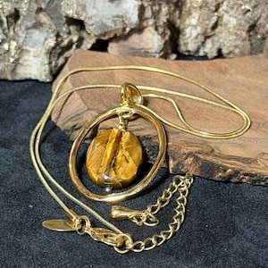 May include: A gold-toned necklace featuring a thin snake chain and a pendant composed of a circular gold-toned frame surrounding a polished, oval-shaped brown and gold tiger's eye gemstone. The necklace includes an adjustable chain extension.