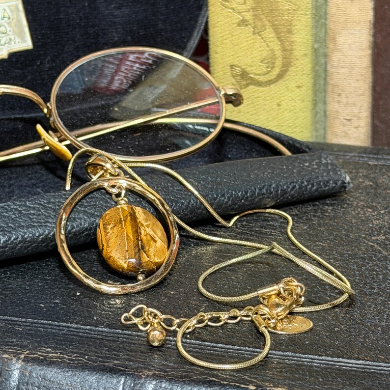 May include: A gold-toned necklace featuring a circular pendant frame containing a polished, brown and gold tiger's eye gemstone. The necklace includes a thin snake chain with an adjustable extender link and a small circular charm at the end.