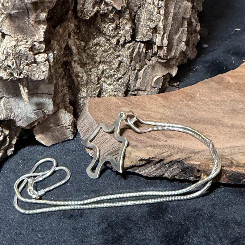 May include: A silver-toned pendant necklace featuring an abstract, openwork design. The pendant is connected to a delicate, snake chain. The necklace rests on a piece of wood with a dark background.