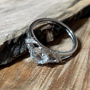 May include: A silver ring with a large, clear, round gemstone as the focal point. The band features smaller accent stones and decorative detailing. The ring is set against a wooden surface, highlighting its design.