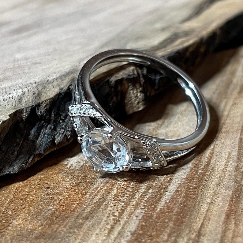 May include: A silver ring with a large, clear, round gemstone as the focal point. The band features smaller accent stones and decorative detailing. The ring is set against a wooden surface, highlighting its design.