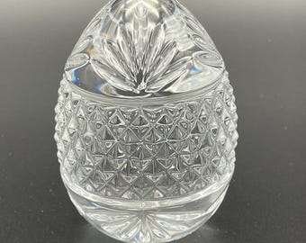 May include: A clear, crystal egg-shaped container with a lid. The lid features a fan-like design, while the body has a diamond-cut pattern. The container is likely made of glass and sits on a dark surface.
