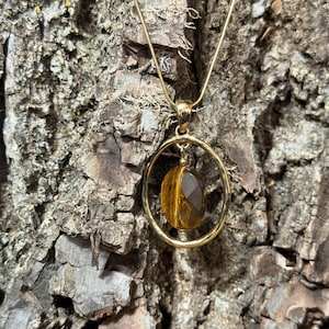 May include: A gold-toned necklace featuring a circular pendant frame that holds a single, faceted, oval-shaped tiger's eye gemstone. The pendant hangs from a thin, matching gold-toned chain, showcasing the natural brown and golden striped patterns.