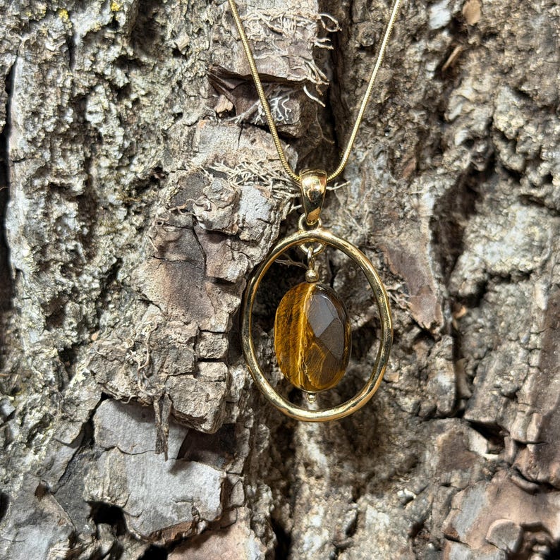 May include: A gold-toned necklace featuring a circular pendant frame that holds a single, faceted, oval-shaped tiger's eye gemstone. The pendant hangs from a thin, matching gold-toned chain, showcasing the natural brown and golden striped patterns.