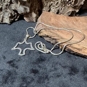 May include: A silver-toned necklace with a pendant shaped like a stylized bird or dove. The pendant is open in the center. The chain is a thin, snake chain style, and the clasp is visible. The necklace is displayed on a dark surface.