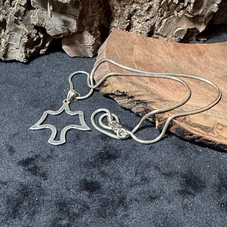 May include: A silver-toned necklace with a pendant shaped like a stylized bird or dove. The pendant is open in the center. The chain is a thin, snake chain style, and the clasp is visible. The necklace is displayed on a dark surface.