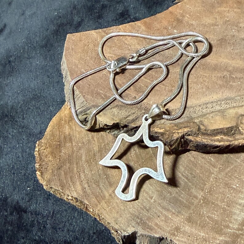 May include: A silver-colored necklace with a pendant in the shape of a stylized bird or leaf. The pendant is open in the center. The necklace is on a wooden surface.