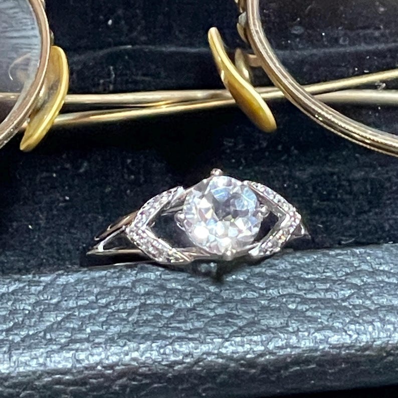 May include: A silver ring with a large, round, clear gemstone at the center. The ring has a decorative band with smaller, clear stones. Gold-rimmed glasses are visible in the background.