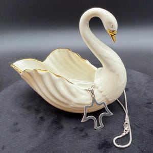 May include: A cream-colored swan-shaped ceramic dish with gold trim, and a silver pendant necklace. The swan has a long, curved neck and a gold beak. The pendant is a stylized dove shape.