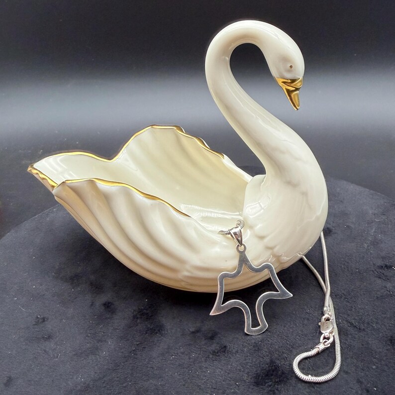 May include: A cream-colored swan-shaped ceramic dish with gold trim, and a silver pendant necklace. The swan has a long, curved neck and a gold beak. The pendant is a stylized dove shape.