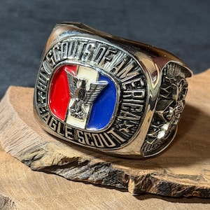 May include: A silver-toned Eagle Scout ring. The ring features a central emblem with red, white, and blue sections, and an eagle. The words "SCOUTS OF AMERICA" and "EAGLE SCOUT" are inscribed around the emblem.