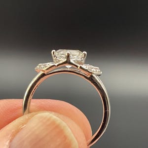 May include: A silver ring with a large, clear, round-cut center stone and smaller accent stones. The ring's band is thin and polished, and the setting has a classic, elegant design. The ring is held up against a dark background.