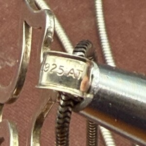 May include: Close-up of a silver necklace clasp with the text "925 AT" stamped on it. The clasp is connected to a silver snake chain. The background is a blurred, reddish-brown color.