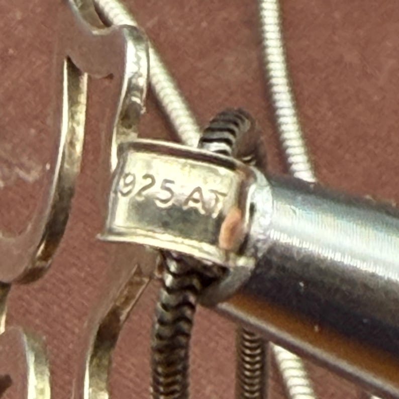 May include: Close-up of a silver necklace clasp with the text "925 AT" stamped on it. The clasp is connected to a silver snake chain. The background is a blurred, reddish-brown color.